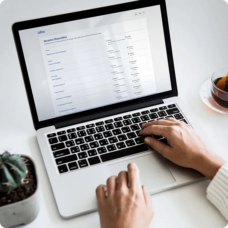 Persona usando una laptop que muestra una página de vacantes de trabajo con una taza de té y una pequeña planta cercana.